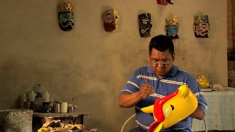 Don Gorgonio pinta con un pincel delgado una máscara amarilla de cresta roja; detrás, en la pared del taller, cuelgan máscaras de reyes y rostros barbados