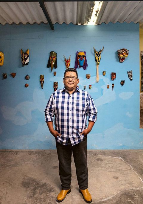 Don Gorgonio de pie en su taller, frente a una pared azul de la que cuelgan sus máscaras de animales y de rostros