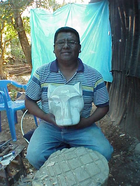 Don Gorgonio sentado al aire libre sostiene una cabeza de felino recién tallada, todavía sin pintar; a sus pies hay un tronco y virutas de madera