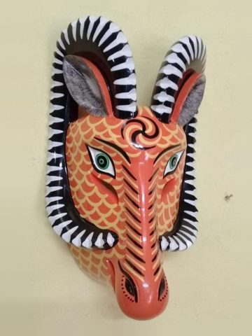 Máscara de borrego anaranjada con escamas amarillas pintadas, espiral negra en la frente y cuernos curvos negros y blancos