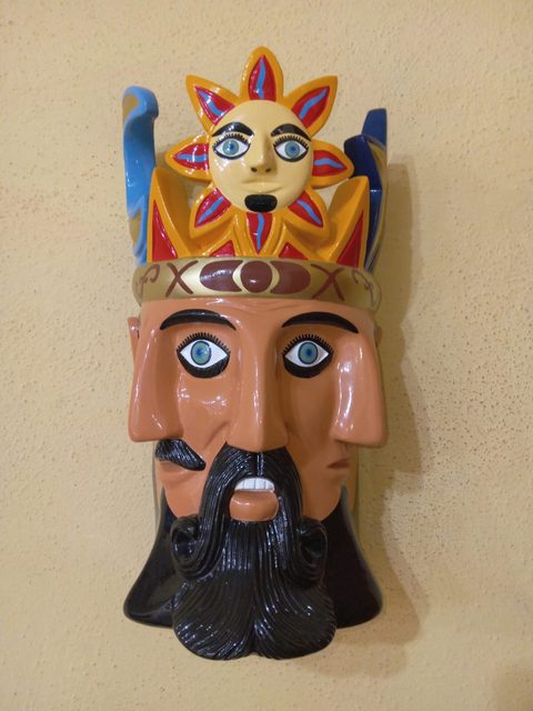 Máscara de El Tiempo: tres rostros color terracota con barba negra rizada, banda dorada y corona rematada por un sol con cara